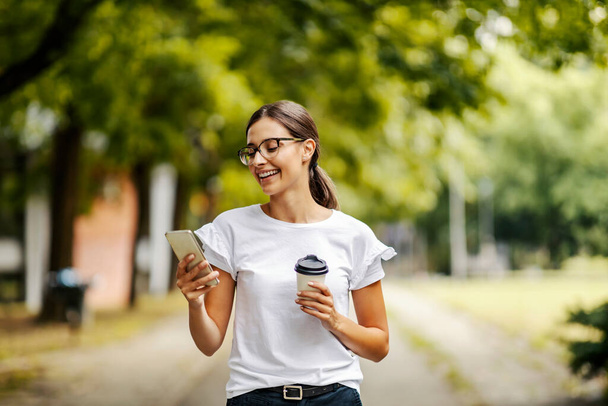 Üniversiteli bir kız kampüste yürüyor ve telefonda önemli bir mesaj alıyor. Ders aralarında kahve molası veriyor, o yüzden elinde tek kullanımlık bir kahve tutuyor.. - Fotoğraf, Görsel