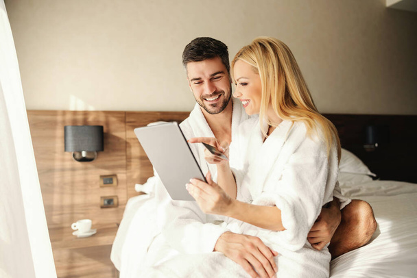 Romantic couple weekend. Sunny morning in a hotel room, a man and a woman making an online reservation in the comfort of a bathrobe. A woman holding a payment card and a tablet in the hugs of men - 写真・画像
