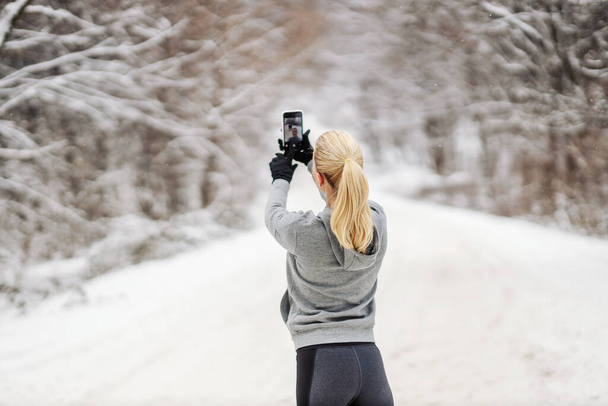 Tylny widok sportowca stojącego w przyrodzie w śnieżny zimowy dzień i robiącego selfie. Technologia, trendy, fitness zimowy - Zdjęcie, obraz