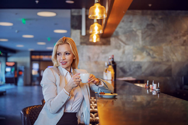 A beautiful middle-aged businesswoman sitting in a hotel cafe and enjoying her coffee. She waiting for a business meeting. Free time, enjoyment, respite, lifestyle - Фото, зображення