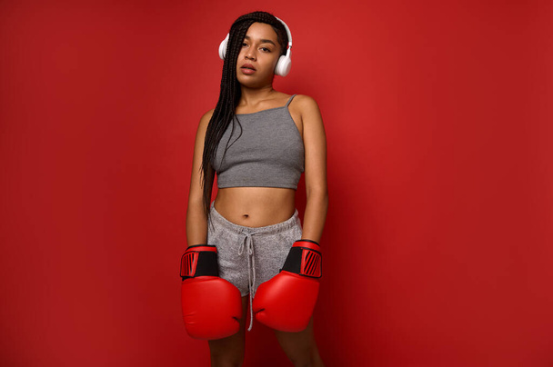 Exhausted young athlete woman boxer, sportswoman wearing wireless headphones, looks at camera posing with red boxing gloves against colored background with copy space. Contact martial art concept - Photo, Image