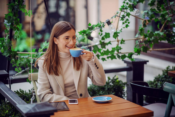 笑顔の女の子がカフェの屋外に座ってコーヒーをすすります。テーブルの上にタブレットと電話があります。ミレニアル世代は、互いに接触する多くの方法があります. - 写真・画像