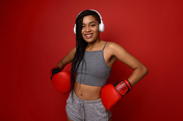Young athlete woman boxer in wireless headphones and hands in boxing gloves on her waist, smiles toothy smile looking at camera. Contact martial art concept on red background with copy space for ad - 写真・画像