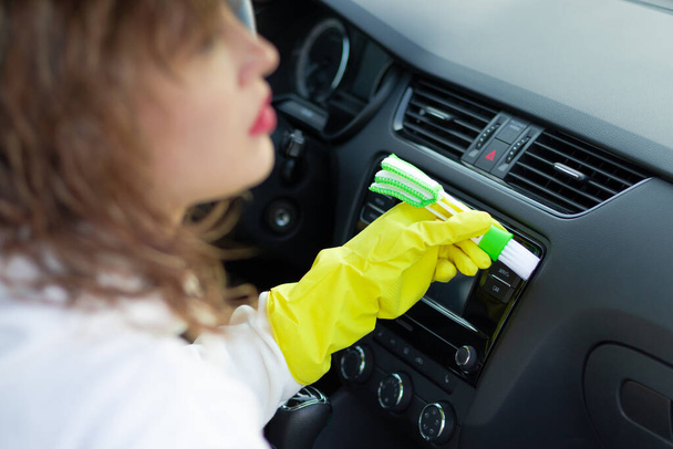 Девушка в жёлтых резиновых перчатках вытирает приборную панель автомобиля из пыли специальной щеткой в яркий осенний день. Выборочный фокус. Крупный план - Фото, изображение