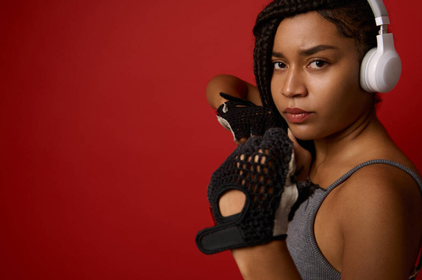Skoncentrowany młody afrykański atletyczna kobieta bokser w słuchawki i czerwone rękawice bokserskie, patrząc na aparat podejmowania proste kopnięcie, izolowane na tle koloru z miejsca kopiowania. Kontakt koncepcja sztuki walki - Zdjęcie, obraz