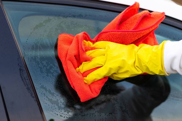 Ręka w żółtej gumowej rękawiczce wyciera szkło samochodu czerwoną szmatą z mikrofibry z brudu w ciepły jesienny dzień. Mokre sprzątanie. Wybiórcze skupienie. Zbliżenie - Zdjęcie, obraz