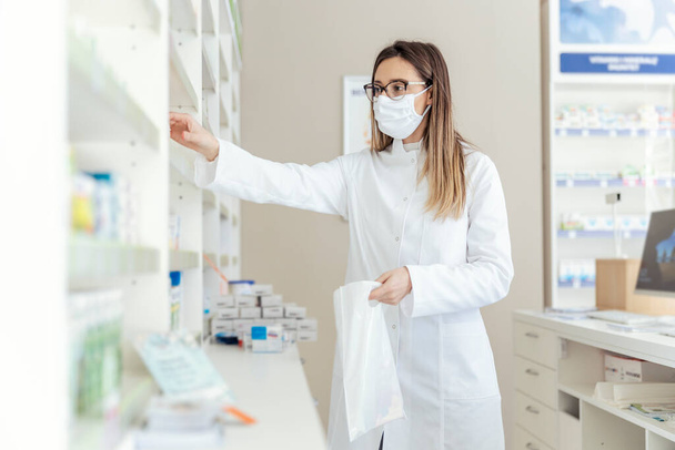 Work in a pharmacy. Packing medicines in a bag. Female employed as a pharmacist and dressed in a white uniform with a protective mask on her face in bags packing boxes of medicines and supplements - Photo, Image