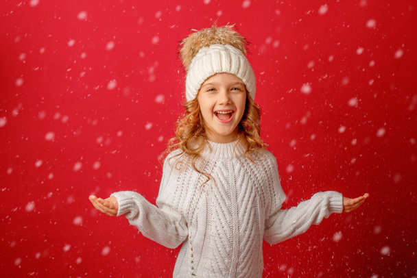 chica en un sombrero de punto coge la nieve sobre un fondo rojo - Foto, imagen