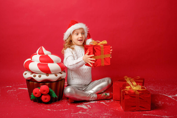 little girl in a Santa hat on a red background with a cake and present - Photo, Image
