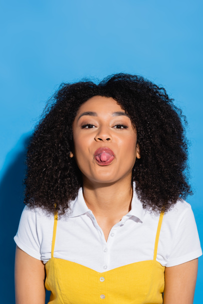 african american woman looking at camera and sticking out tongue on blue - Fotoğraf, Görsel
