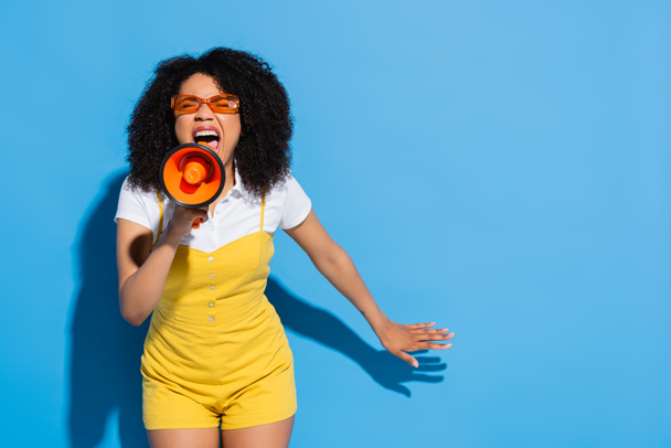 african american woman in orange eyeglasses screaming in megaphone on blue - Foto, immagini