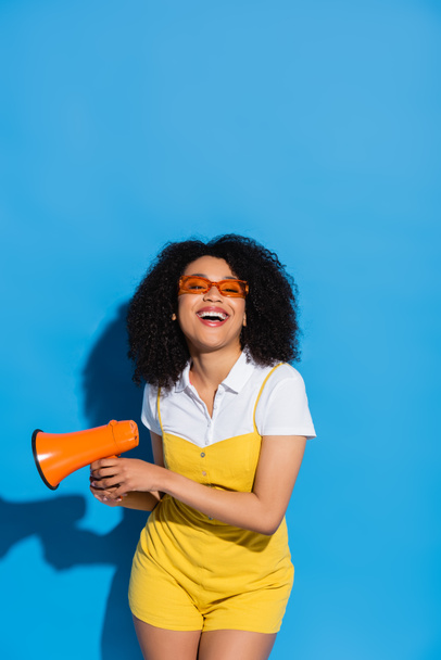 cheerful african american woman in trendy eyeglasses holding megaphone on blue - Foto, immagini