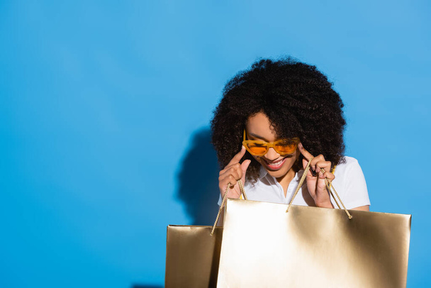 Χαρούμενη Αφροαμερικανή γυναίκα με χρυσές τσάντες για ψώνια που προσαρμόζει κίτρινα γυαλιά στο μπλε - Φωτογραφία, εικόνα