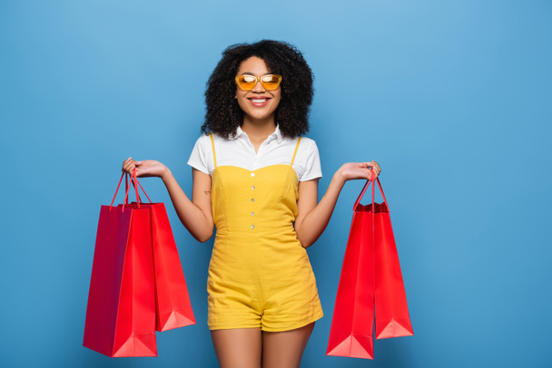 happy african american woman in yellow eyeglasses and jumpsuit posing with shopping bags on blue - Zdjęcie, obraz