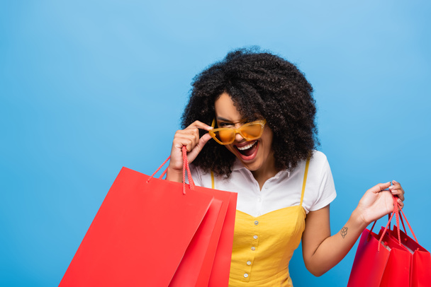 excited african american woman with shopping bags touching trendy eyeglasses isolated on blue - Valokuva, kuva