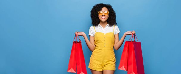 cheerful african american woman in yellow jumpsuit and eyeglasses standing with shopping bags isolated on blue, banner - Foto, Bild