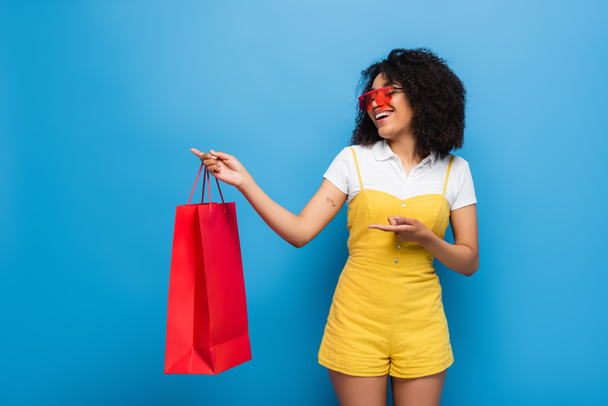 Веселая африканская американка в красных очках и жёлтом комбинезоне, указывающая на коралловую сумку для покупок на синем - Фото, изображение