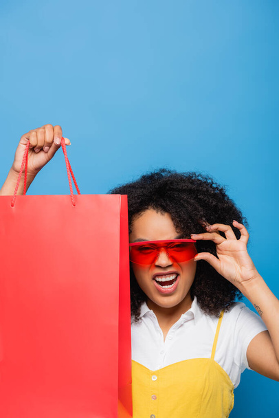 thrilled african american woman with shopping bag adjusting trendy eyeglasses isolated on blue - Foto, imagen