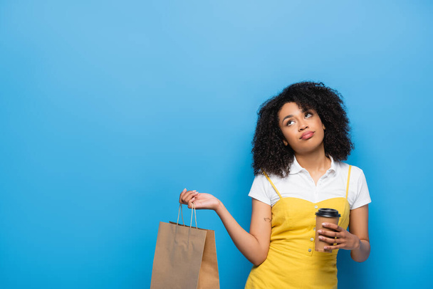 displeased african american woman with paper cup and shopping bag looking up on blue - Foto, immagini