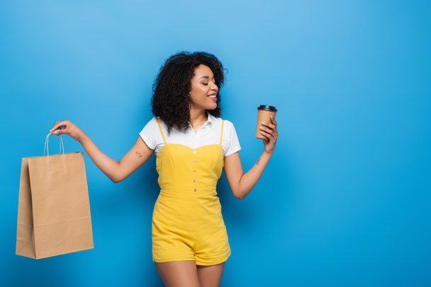 donna afro-americana sorridente che tiene drink da asporto e shopping bag su blu - Foto, immagini