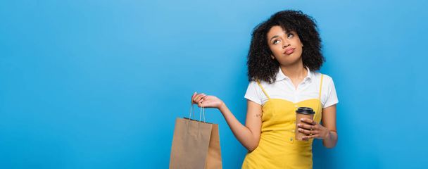 mujer afroamericana molesta con café para ir y bolsa de compras mirando hacia arriba en azul, pancarta - Foto, imagen