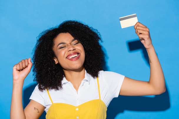 興奮したアフリカ系アメリカ人女性目を閉じてクレジットカードを保持し、青で勝つジェスチャーを示す - 写真・画像