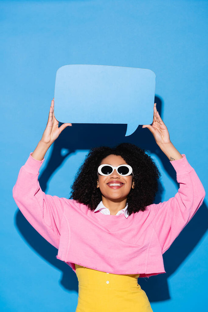 wesoła Afroamerykanka w stylowych okularach przeciwsłonecznych z pustą bańką mowy na niebiesko - Zdjęcie, obraz