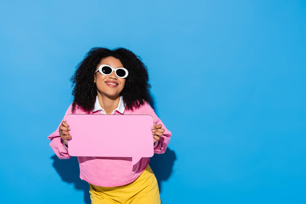joyful african american woman in trendy sunglasses and pink sweatshirt holding blank speech bubble on blue - 写真・画像