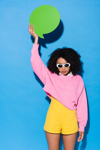 mujer afroamericana en gafas de sol con estilo sosteniendo burbuja de pensamiento vacío en la mano levantada en azul - Foto, imagen