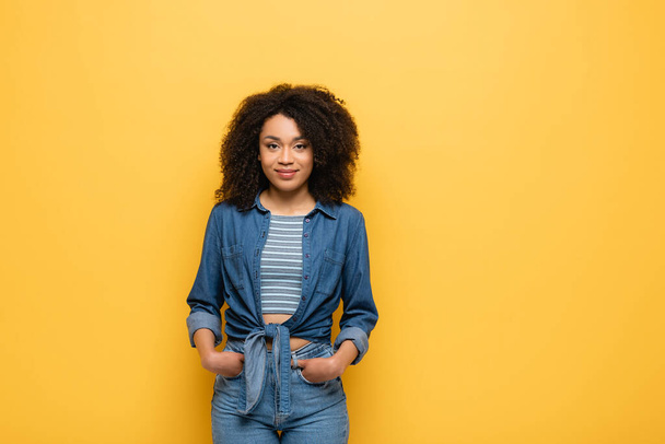 smiling african american woman in denim shirt posing with hands in pockets of jeans isolated on yellow - Фото, зображення