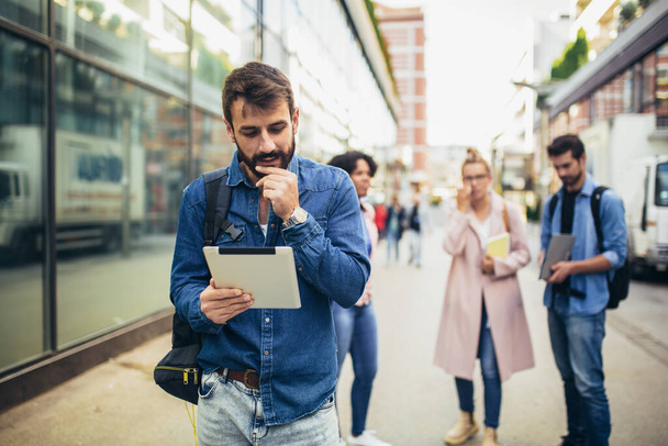 Giovane uomo in possesso di tablet digitale mentre in piedi sullo sfondo di università e amici. - Foto, immagini
