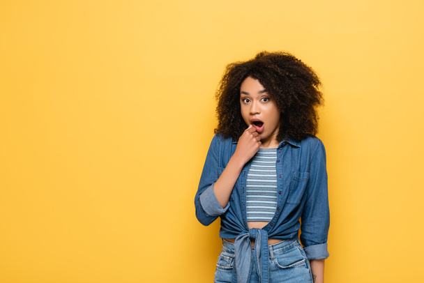 shocked african american woman in denim short looking at camera isolated on yellow - Photo, Image
