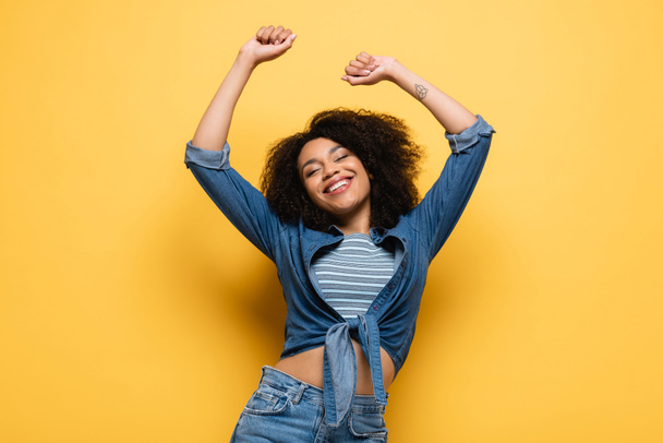 joyful african american woman in denim shirt dancing with closed eyes and raised hands on yellow - Foto, immagini