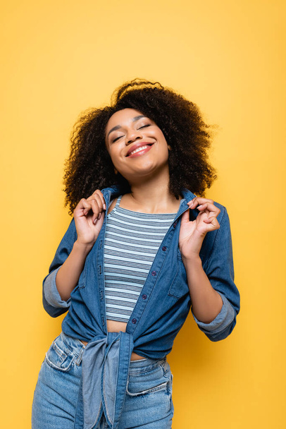 pleased african american woman touching denim shirt while posing with closed eyes isolated on yellow - Фото, изображение