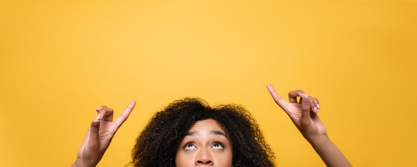 partial view of african american woman pointing with fingers while looking up isolated on yellow, banner - Foto, afbeelding