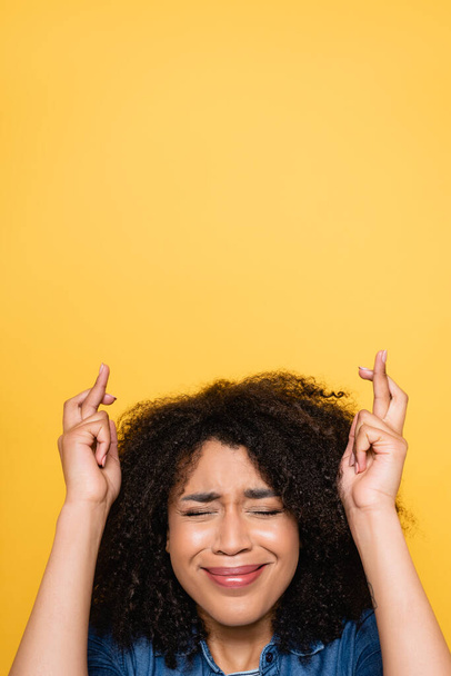 worried african american woman with closed eyes holding crossed fingers isolated on yellow - Photo, Image