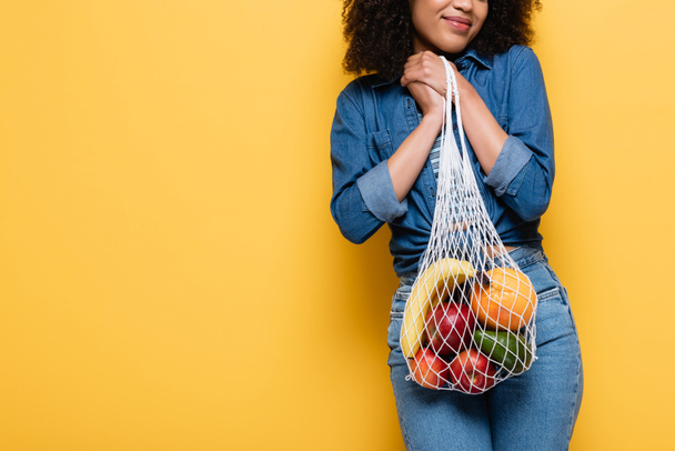 cropped view of smiling african american woman holding ripe fruits in string bag on yellow - Photo, Image