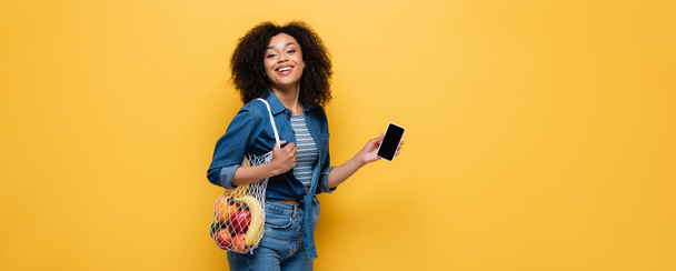mujer afroamericana feliz sosteniendo teléfono inteligente y bolsa de cuerda con frutas aisladas en amarillo, pancarta - Foto, Imagen