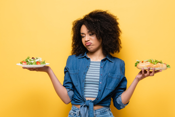 displeased african american woman looking at vegetable salad while holding hot dog isolated on yellow - Foto, afbeelding