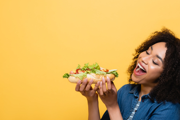 pleased african american woman eating tasty hot dog isolated on yellow - Fotografie, Obrázek