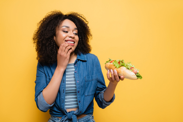 freudige afrikanisch-amerikanische Frau beim Anblick köstlicher Hot Dogs isoliert auf gelb - Foto, Bild