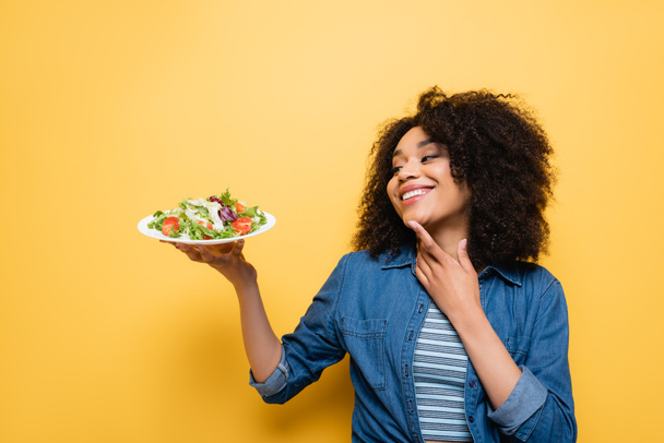 pleased african american woman in denim shirt holding fresh vegetable salad on yellow - Foto, afbeelding