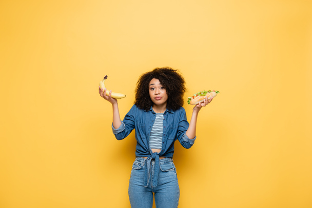 confused african american woman looking at camera while choosing between bananas  and hot dog on yellow - Φωτογραφία, εικόνα