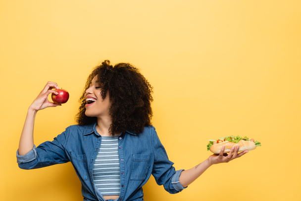 pleased african american woman biting fresh apple while holding hot dog on yellow - Zdjęcie, obraz