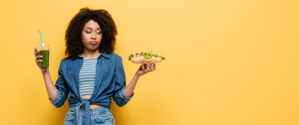 confused african american woman looking at hot dog while holding fresh smoothie on yellow, banner - Photo, Image