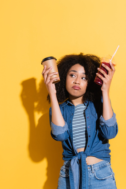mujer afroamericana confusa elegir entre café y batido fresco en amarillo - Foto, Imagen