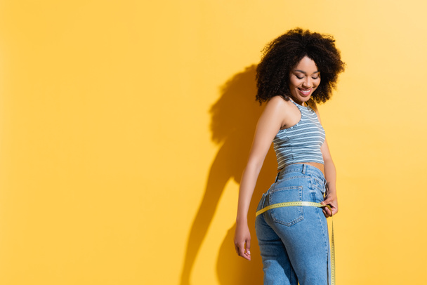 slim and joyful african american woman in jeans and tank top measuring hips on yellow - Фото, зображення