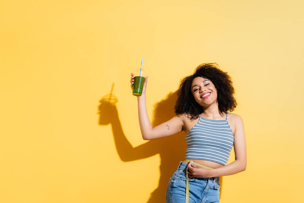 slim and happy african american woman with fresh smoothie measuring waist on yellow - Foto, Imagem