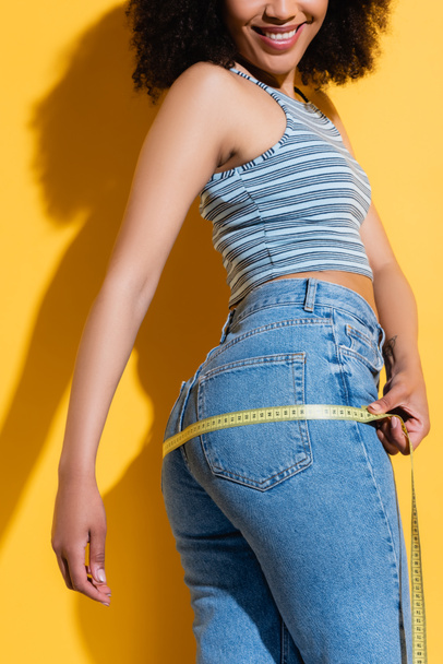 cropped view of slim african american woman in jeans measuring hips on yellow - Fotó, kép