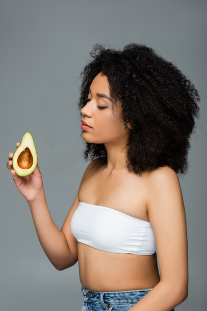 кучерява афроамериканська жінка в білому верхньому тримає половину стиглого авокадо ізольовано на сірому
 - Фото, зображення